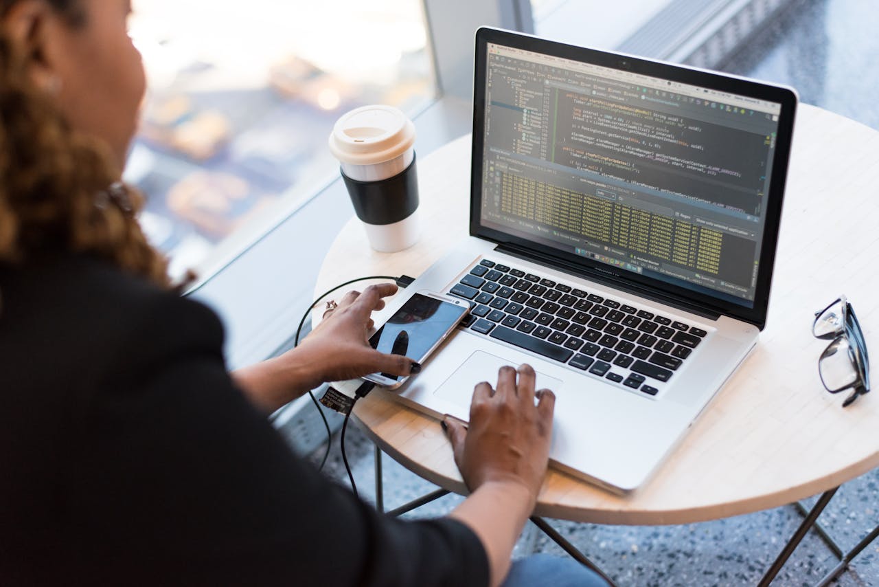 Services Black woman programming on a laptop with coffee, smartphone, and glasses on a desk in an office.